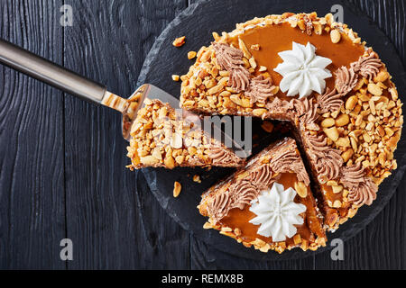 Erdnuss Kuchen gekrönt mit Kakao Peanut Butter, Sahne und Eischnee Rosen in Scheiben geschnitten auf einer Steinplatte auf einen hölzernen Tisch, Blick von oben, Nahaufnahme, Fl Stockfoto