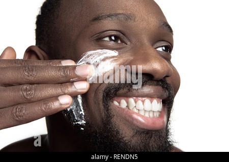 Junge afrikanische amerikanische Kerl Anwendung Gesicht Creme unter den Augen auf weißem Hintergrund. Portrait einer jungen Glücklich lächelnde afrikanischer Mann im Studio. High Fashion männliches Modell. Stockfoto