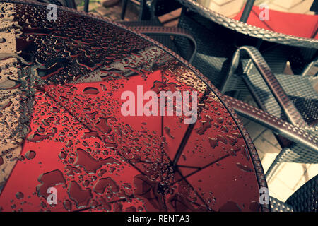 Glas Tisch in einem Sommer Cafe mit Wassertropfen und Reflexion eines roten Regenschirm neben Metall Korbstühlen Stockfoto