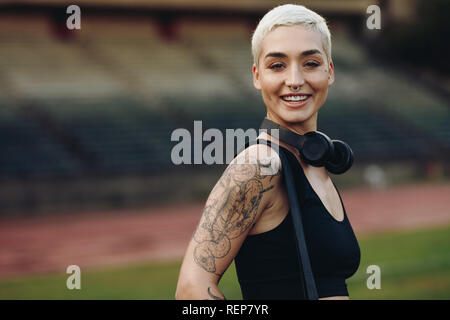Freundliche fitness Frau in Leichtathletikstadien mit ihrer Tasche und kabellosen Kopfhörern. Seitenansicht eines weiblichen Athleten in einer Schiene und Feld st Stockfoto