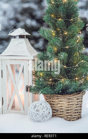 Weiße hölzerne Laterne mit weißem Wachs Kerze angezündet und kleine Weihnachten Tannenbaum mit Micro-Led leuchtet in Braun Rattan Blumentopf im Schnee, verschneite Tannen Stockfoto