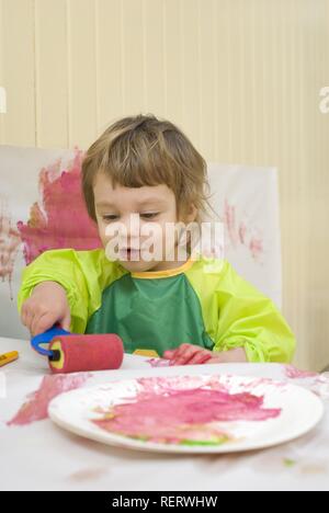 Ein zwei Jahres altes Mädchen in einem Kunststoff-smock Malerei in ein Spielzimmer mit einem Pinsel und Rolle Stockfoto