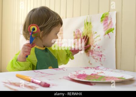 Ein zwei Jahres altes Mädchen in einem Kunststoff-smock Malerei in ein Spielzimmer mit einem Pinsel und Rolle Stockfoto