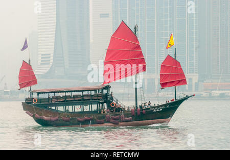 Segelboot mit rotem Segel und Gelb und Lila Fahnen, mit Menschen an Bord, die in der Ferne den Hafen von Hong Kong, China. Stockfoto