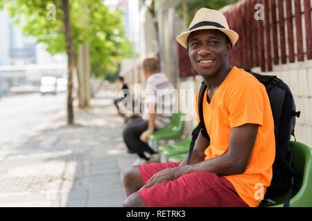 Junge glücklich schwarzen Afrikanischen touristische Mann an der Bushaltestelle sitzen Stockfoto