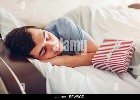 Junger Mann schlafen im Bett in der Nähe Geschenkbox von seiner Frau für Valentines Tag. Überraschung für Urlaub Stockfoto