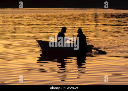 Ein Mann und eine Frau in einem kleinen Ruderboot sind gegen die sanft wellige Wasser im goldenen Abendlicht. Stockfoto