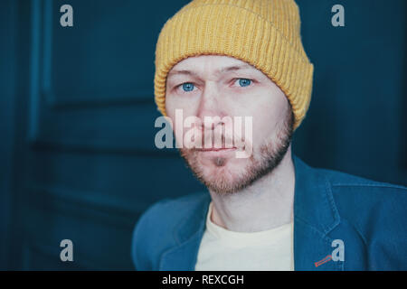 Attraktive nach bärtigen Mann Hipster in gelben Hut misstrauisch an Kamera auf der blauen Wand Hintergrund Stockfoto