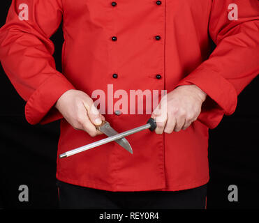 Koch in roter Uniform schärft ein Messer, schwarzer Hintergrund Stockfoto