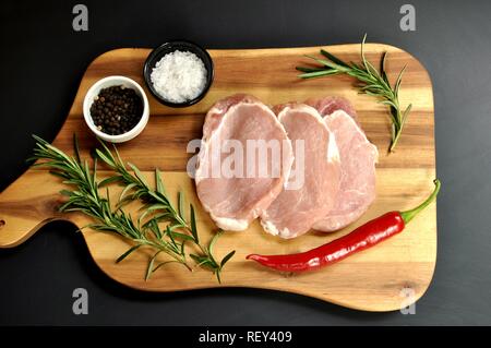 Roh frisch ungekocht geschnittenes Schweinefleisch Filetgericht mit Rosmarin, Pfeffer, Salz, rote Chili Pfeffer auf Holzbrett und schwarzem Hintergrund. Tisch auf der obersten Platte. Stockfoto