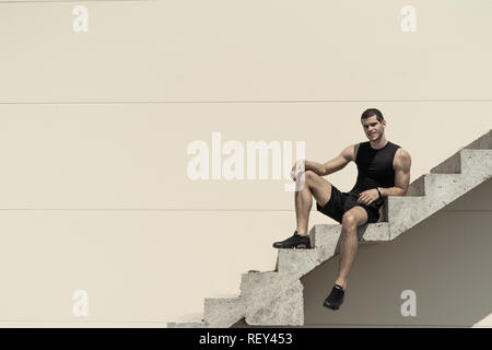 Sportlich unbeschwerte Sportsman model posing beim Sitzen und Lächeln in entspannter Pose auf Treppen Platz Kopieren im Hintergrund. Umsatz, Fitnessclubs, Adv Stockfoto