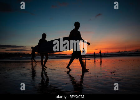 Silhouette von Surfern, die im Sonnenuntergang mit einem Longboard spazieren. Tamarindo Beach, Tamarindo, Costa Rica, Mittelamerika Stockfoto