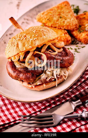 Leckere deutsche Bratwurst Burger mit würzig gegrilltem Schweinefleisch Wurst und Zwiebel Ringe auf einem Sesambrötchen, serviert auf einem Teller mit Serviette und Utensilien Stockfoto