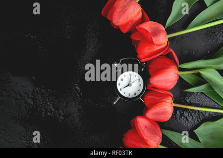 Schwarz Wecker in der Nähe von Bouquet rote Tulpen auf schwarzem Hintergrund. Mutter oder Frau Tag. Grußkarte. Guten Morgen Frühstück. Kopieren Sie Platz. Feder. Stockfoto