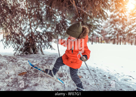 Die ersten Schritte eines Junge Kind 3-5 Jahre alt, auf der Kinder Ski mit Stöcken. Im Park im Winter. Das Konzept der Sport- und Freizeitaktivitäten von einem jungen Alter. Klettert in den Schnee. Stockfoto