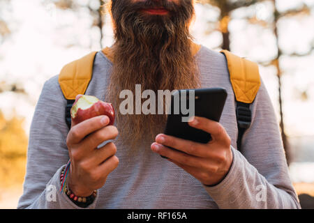 Nahaufnahme des bärtigen Mann mit Handy und essen einen Apfel auf eine Wanderung im Wald Stockfoto