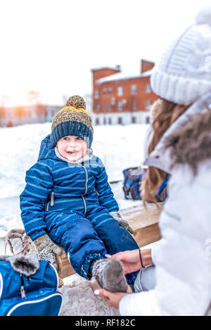 Mom pereobuvaet Sohn sitzen auf einer Bank in Winter Park. Pflege einer jungen Mutter für ein Kind ist ein Junge. Trägt Socken entfernen Skates änderungen Schuhe. Stockfoto