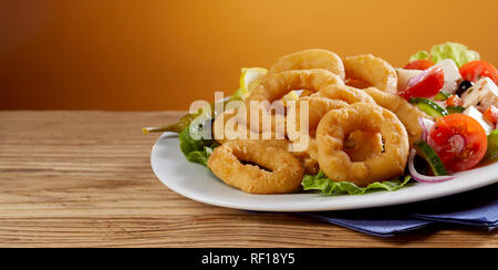 Golden gebratene Calamari Ringe mit frischen griechischen Salat auf einen Teller auf einem hölzernen Zähler mit Kopie Raum in Banner Format serviert. Stockfoto