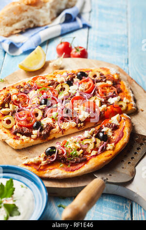 Leckere griechische Pizza mit Oliven, Schafskäse, Tomaten und Zwiebeln auf einem knusprig dünne Basis mit raita an der Seite auf einer hölzernen Schneidebrett in Scheiben geschnitten serviert. Stockfoto