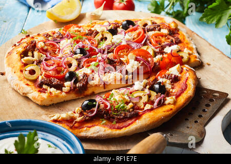 Leckere griechische Pizza auf einem dünnen Teig Base in Scheiben geschnitten und serviert mit einem Spachtel auf einem Holzbrett mit einer Schüssel mit raita Joghurt Dressing zur Seite Stockfoto