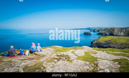 Leute sitzen auf den Felsen von Tintagel Insel mit Blick auf das Meer, Klippen. Ruinen einer Mauer der Burg Tintagel des legendären König Artus in Cornwall, Großbritannien Stockfoto