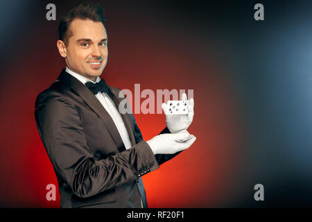 Berufliche Tätigkeit. Magier in Anzug und Handschuhe isoliert auf Wand zeigen Trick scharren Karten lächelnd verspielten stehend Stockfoto