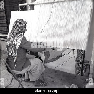 1950 s, historischen, einen Araber in traditionelle arabische Kleidung mit Kopftuch sitzt auf einem kleinen Stuhl mit einer Hand Webstuhl einen dekorativen Teppich zu weben, Saudi-Arabien. Stockfoto