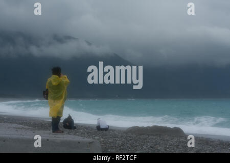 Frau in Regenmantel Bilder in den regnerischen, windigen Wetter, azurblauen Wasser waschen über Kieselstrand, zentrale Bergkette im Hintergrund, Hualien, Taiwan Stockfoto