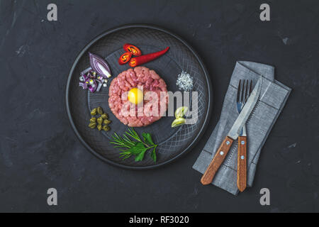 Steak Tartar mit Eigelb und Zutaten aus schwarzer Keramik Teller, Besteck Messer und Gabel auf schwarzen Stein Beton texturierte Oberfläche Hintergrund. Cop Stockfoto