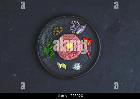 Steak Tartar mit Eigelb und Zutaten aus schwarzer Keramik Teller, Besteck Messer und Gabel auf schwarzen Stein Beton texturierte Oberfläche Hintergrund. Cop Stockfoto
