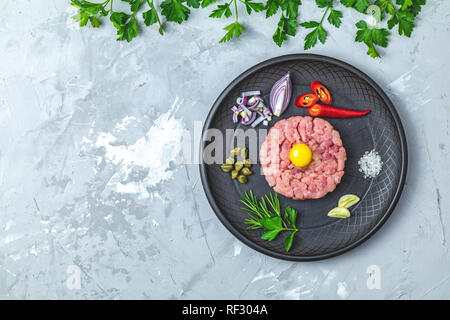 Steak Tartar mit Eigelb und Zutaten aus schwarzer Keramik Teller, Besteck Messer und Gabel auf hellen grauen Stein Beton texturierte Oberfläche Hintergrund Stockfoto