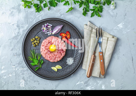 Steak Tartar mit Eigelb und Zutaten aus schwarzer Keramik Teller, Besteck Messer und Gabel auf hellen grauen Stein Beton texturierte Oberfläche Hintergrund Stockfoto