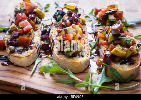 Lecker Tomaten und Oliven Bruschetta mit Kapern, Rucola eine Balsamico beträufeln, serviert auf einem Schneidebrett aus Holz Stockfoto