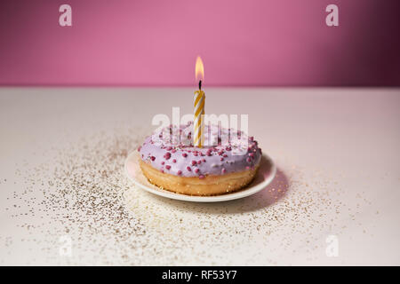 Brennende Kerze in der Mitte der glasierten Krapfen mit funkelt auf weißen Tisch auf rosa Hintergrund Stockfoto