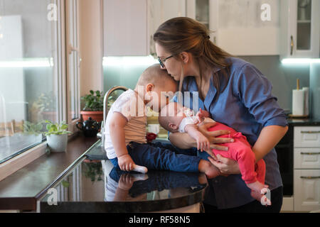 Junge Mutter hält kleine Tochter mit Schnuller und küsse Leiter der älteste Sohn. Junge küsst seine Schwester. Zeit mit der Familie. Stockfoto