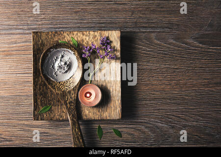 Natürliches Grau Schlamm Maske Creme auf Holzlöffel mit Kerze brennen und Blume Zweig als Dekoration auf Holz- Fach auf Tisch. Flach anzeigen, kopieren. Stockfoto