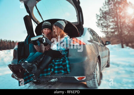 Gerne Freunde im Winter Wald. Zwei Mädchen sitzen im Kofferraum des Autos trinken Kaffee Stockfoto