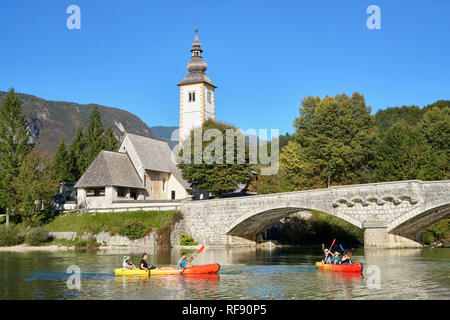 Die Kirche des Hl. Johannes des Täufers, des Cerkev Sv. Janeza Krstnika und Lake Bohinj, Ribcev Laz, Bohinj, Gorenjska, Slowenien. Mit Kanuten auf dem See Stockfoto