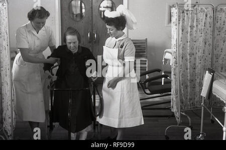1950, historische, zwei weibliche Krankenschwestern helfen eine verletzte Frau Patient in einem Mobilität Stuhl bewegen Sie sich in einem Krankenhaus, England, Großbritannien Stockfoto