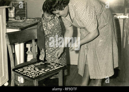 1959, historische, ein junge wearng eine Schürze in einer Küche zu Hause mit seiner Mutter mit einem Fach der kleinen süßen Kekse oder Cookies er gemacht hat. Es wird angenommen, dass die Förderung junger Kinder zu kochen hilft Ihnen ihre Sinne, Fähigkeiten und Selbstvertrauen aufgebaut. Stockfoto