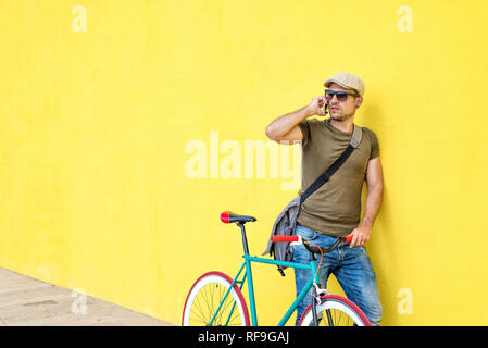 Seitenansicht eines jungen Mannes mit einem Vintage bike und legere Kleidung und Sonnenbrillen gegen eine gelbe Wand, während Sie ein Mobiltelefon in einem Stockfoto