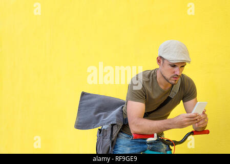 Seitenansicht eines jungen Mannes mit einem Vintage bike und legere Kleidung und Sonnenbrillen gegen eine gelbe Wand, während Sie ein Mobiltelefon in einem Stockfoto