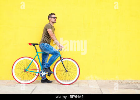 Seitenansicht eines jungen trendigen Mann mit einem festen Bike legere Kleidung, wenn Sie sich nicht gegen eine gelbe Wand im Freien an einem sonnigen Tag Stockfoto
