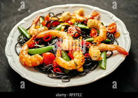 Frische, leckere schwarze Spaghetti mit gegrillten Garnelen, Tomaten, Peperoni, auf einer weißen Platte Stockfoto