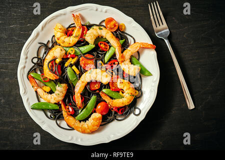 Frische, leckere schwarze Spaghetti mit gegrillten Garnelen, Tomaten, Peperoni, auf einer weißen Platte Stockfoto