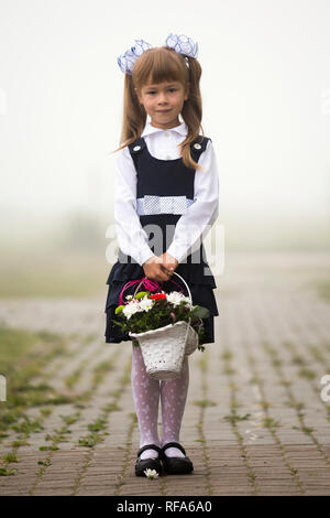 Full-length Portrait von cute adorable erster Sortierer Mädchen in Schuluniform und weißen Bögen mit Blumen Warenkorb Zusammensetzung stehen auf gepflasterten leere Straße Stockfoto