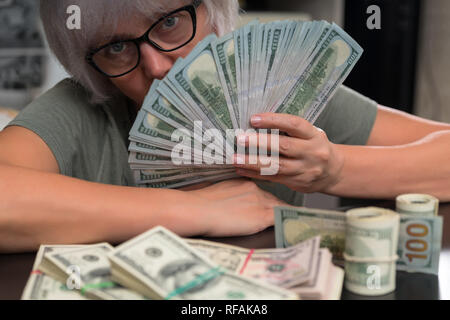 Frau mit einer Handvoll 100 Dollar-Scheine ihr Gesicht verstecken mit einem großen Stapel von Walzen und bündelt vor ihr in einem konzeptionellen finanzielle Bild Stockfoto