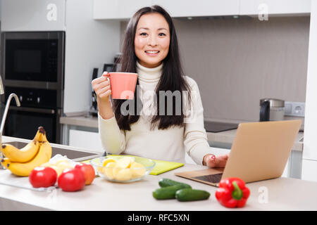 Bild der glücklichen asiatischen Brünette mit Tasse und Laptop am Tisch mit Gemüse und Obst in der Küche Stockfoto