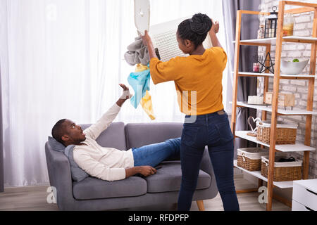 Junge Frau werfen schmutzige Kleidung auf Faul Mann auf dem Sofa liegend Stockfoto