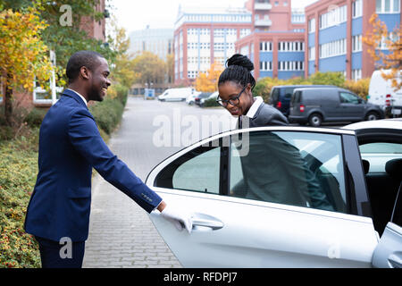 Gerne afrikanische Mann Valet Öffnen der Tür für die Geschäftsfrau, die sich aus einem Auto auf der Straße Stockfoto